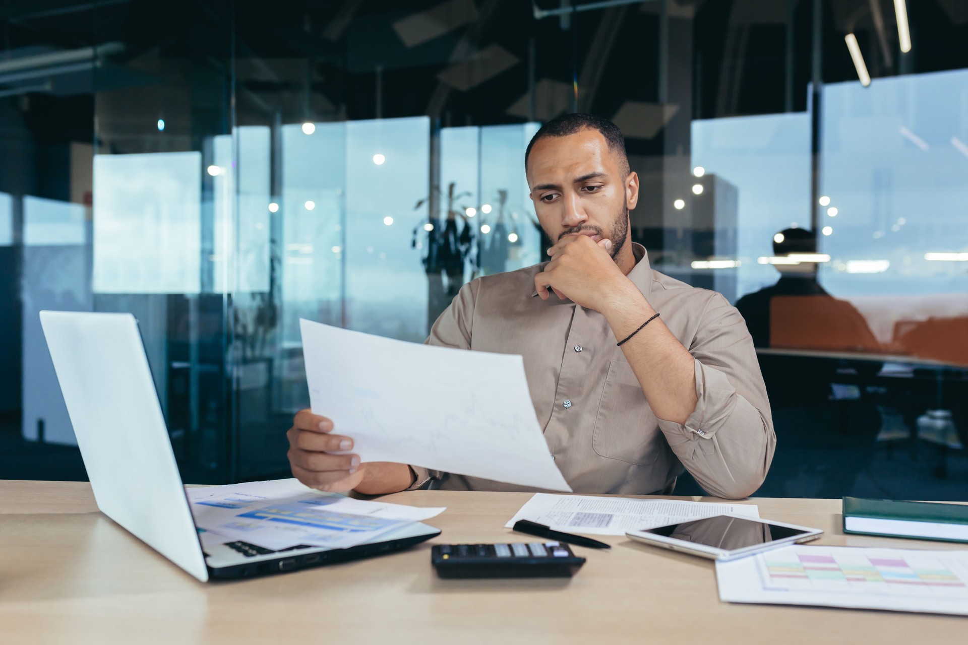 Pensive serious businessman reading financial report, hispanic businessman holding document in hands looking disappointed, working inside modern office with laptop behind paper work